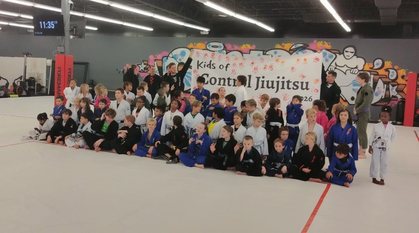 The whole kids program sitting together on the mat in front of the Control Jiu-Jitsu mural